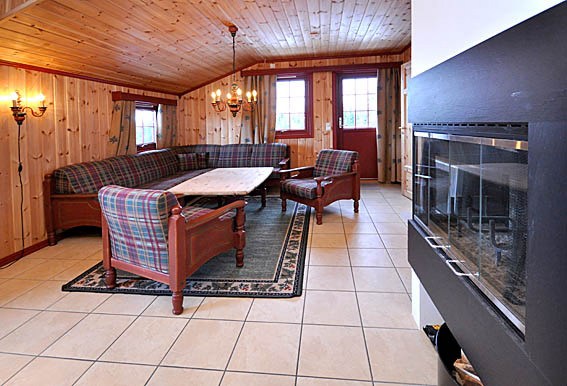 En hyggelig stue i en Wagrain-hytte med trævægge og -loft, med plaid sofaer og stole omkring et sofabord af træ. En pejs er til højre, og en lysekrone hænger ovenfra. Naturligt lys strømmer ind gennem et vindue og en dør, perfekt til afslappende afterski-eventyr.