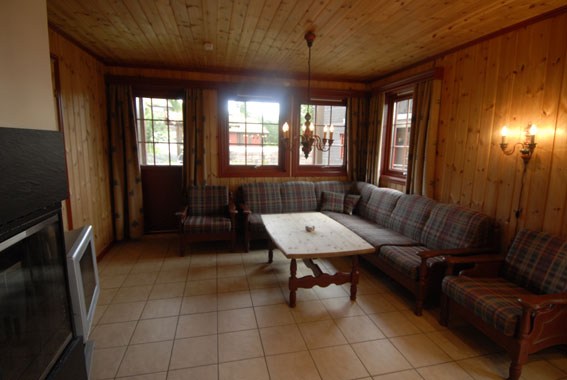 En hyggelig stue med trævægge og -loft, med en stor plaidmønstret sofa, en lænestol, et sofabord i træ og en lysekrone. Stemningen føles som et charmerende tilflugtssted i Saalbach under din sommerferie Østrig, med vinduer og dør, der tillader naturligt lys at oplyse rummet.