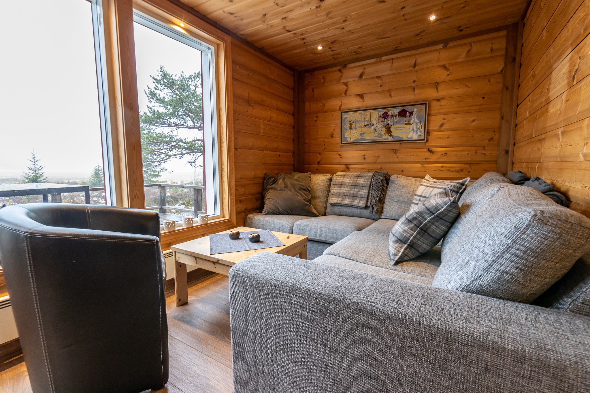 Hyggelig træstue i Westendorf-stil, med en stor grå sektionssofa med plaidpuder og et sofabord i træ. En sort læderstol sidder ved vinduer med udsigt til træer, der minder om skirejsers eventyr. Et indrammet billede pryder væggen, da naturligt lys fylder rummet.