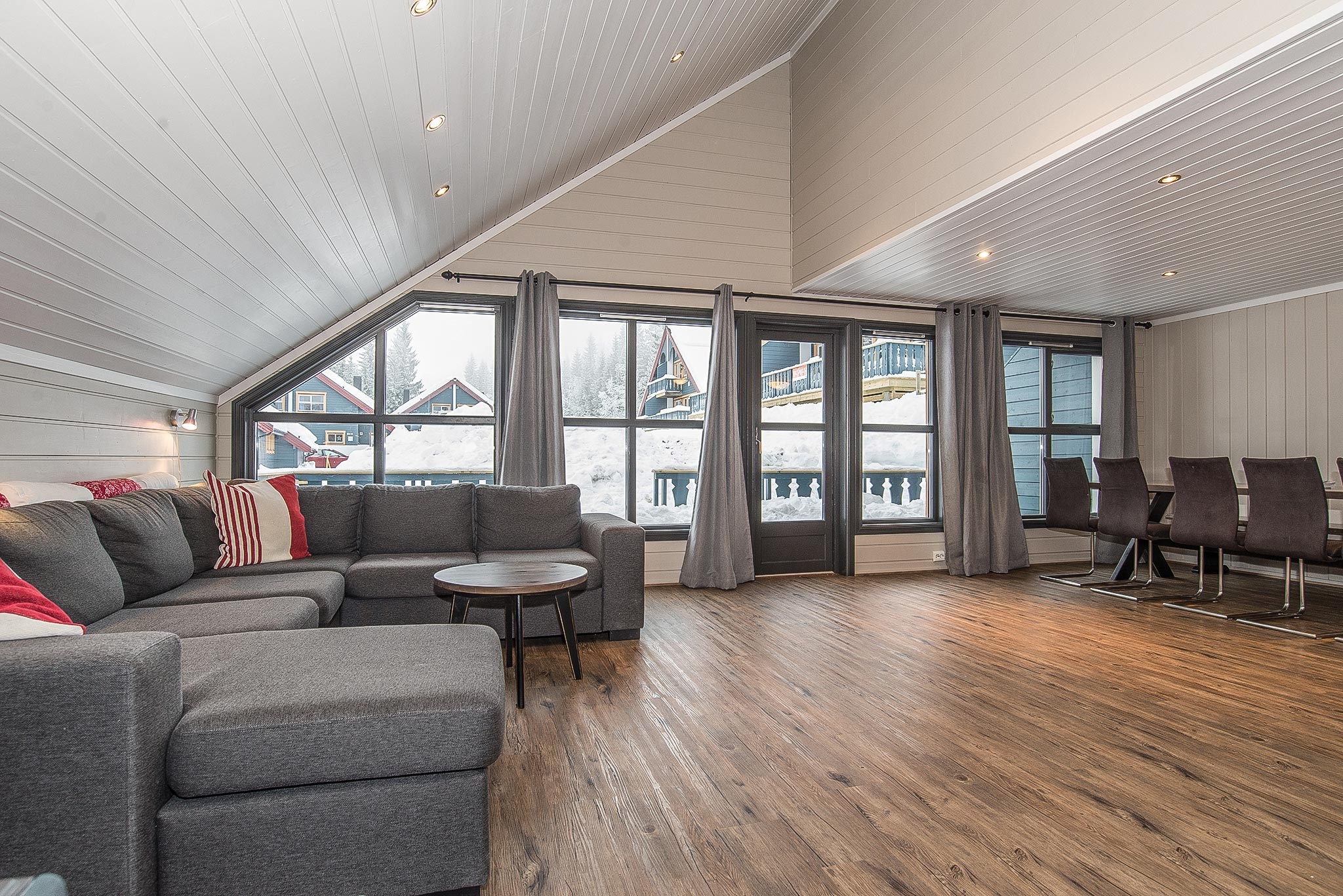 En rummelig stue med hvælvet loft i Westendorf har en grå sektionssofa prydet med røde og hvide puder. Store vinduer har udsigt til et sneklædt landskab og træhytter. Et spisebord med mørke stole er til højre, og lyse trægulve dækker området.