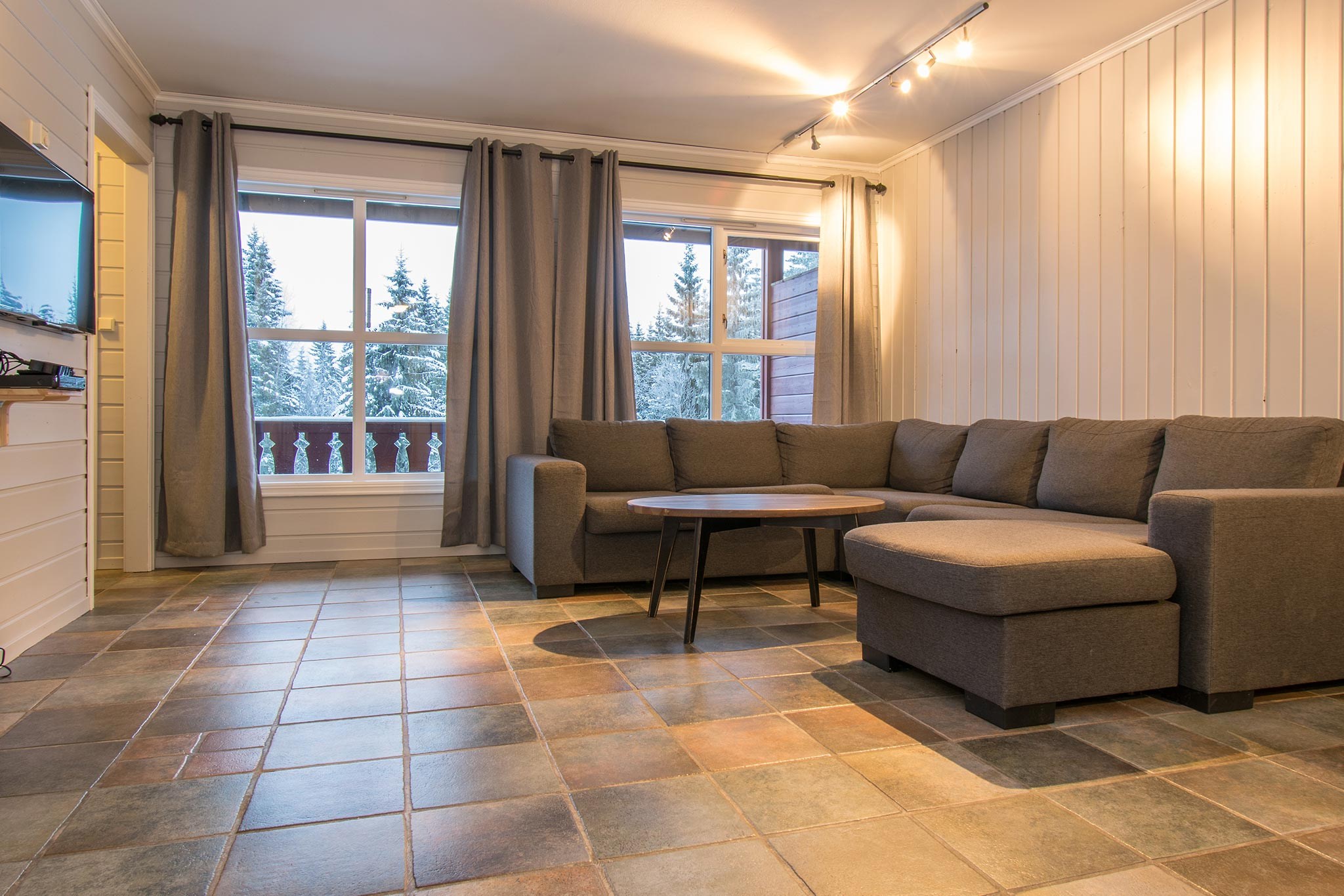 En hyggelig stue perfekt til afterski-afslapning i Westendorf, med en stor grå sektionssofa og et sofabord i træ på et klinkegulv. De hvide panelvægge og store vinduer med gardiner giver udsigt til snedækkede træer udenfor. Sporbelysning sætter den indbydende stemning.