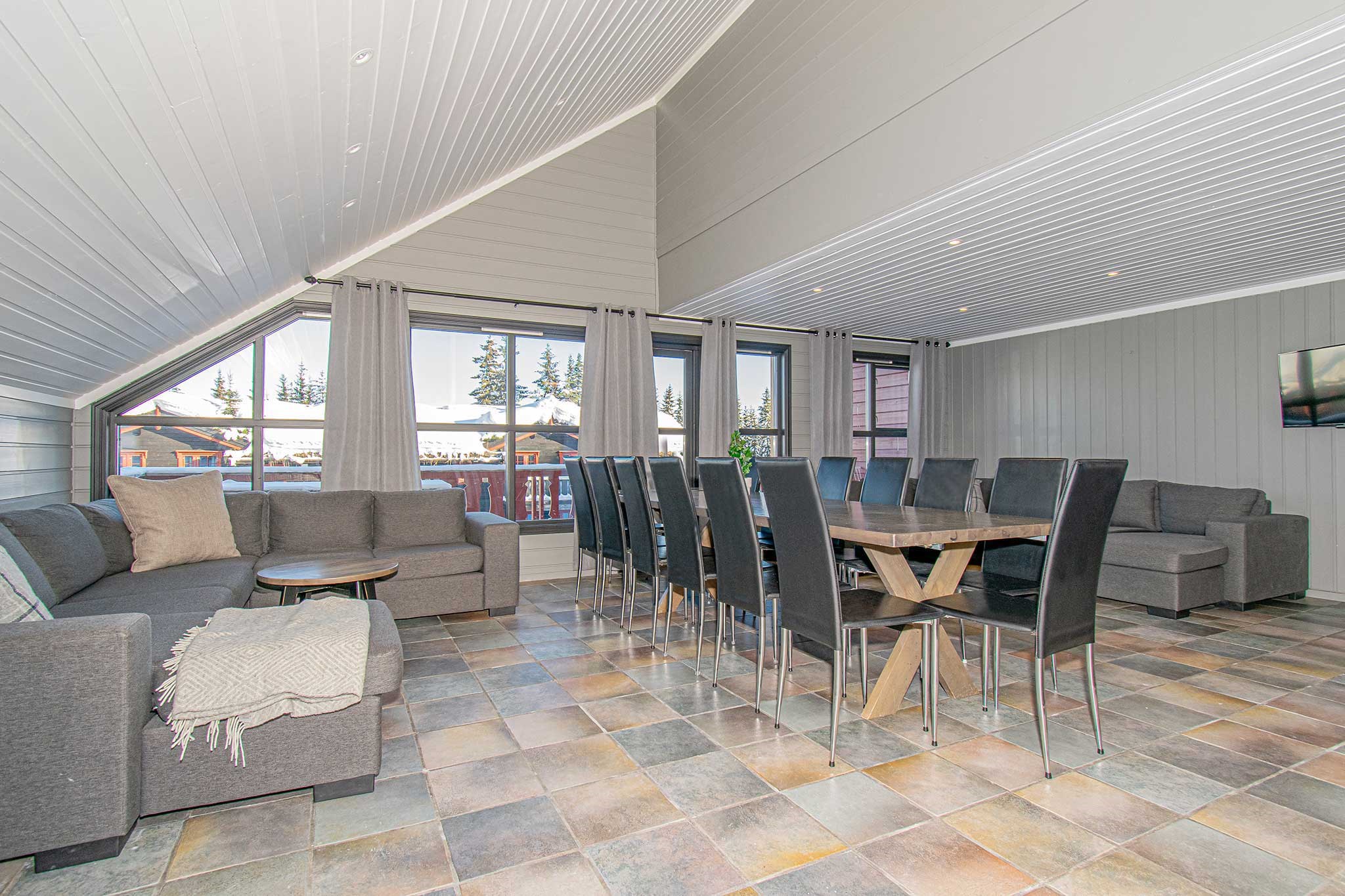 En rummelig, moderne stue med højt, skrå loft har grå sofaer og et langt spisebord med sorte stole. Store vinduer slipper naturligt lys ind og giver en udsigt, der minder om en Westendorf skiferie, med træer og et hus udenfor. Gulvet er flisebelagt i stenmønster.