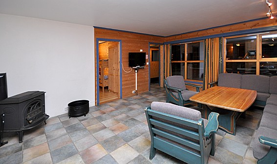 En hyggelig opholdsstue med trævægge og klinkegulv, perfekt til et skirejseretreat. Den har en pejs til venstre, et fladskærms-tv på væggen, et spisebord i træ med stole og et stort vindue med gardiner til højre. En lille gang fører til et andet værelse i Wagrain.