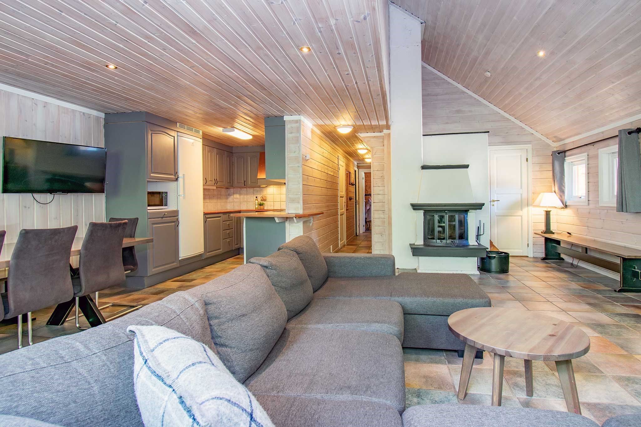 Hyggelig opholdsstue med en moderne L-formet grå sofa, lille rundt træbord og et væghængt tv. Det åbne køkken har et spisebord og stole i baggrunden. Trævægge og -loft giver en varm, rustik følelse, der minder om et fredfyldt sommerferie Østrig-tilflugtssted.
