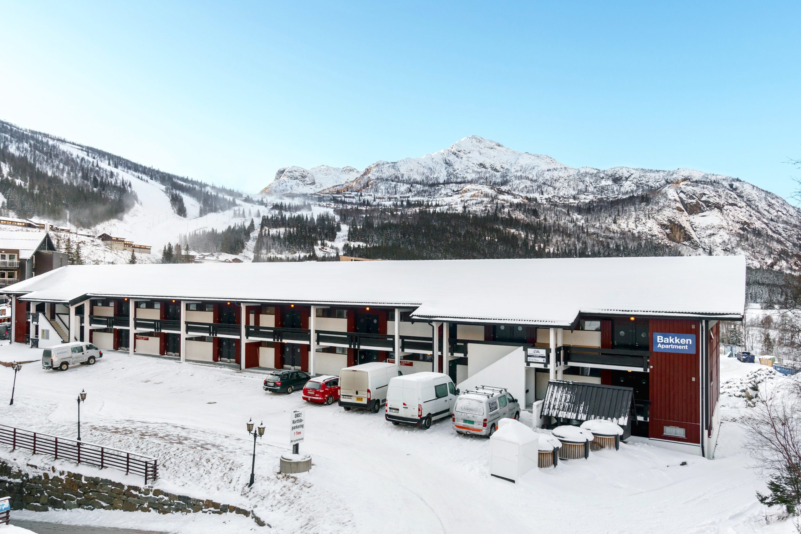 Et snedækket hotel i Westendorf, med flere køretøjer parkeret udenfor, putter sig mod en bjergrig baggrund. Den blå himmel står i kontrast til det hvide landskab og skaber en fredfyldt vinterscene perfekt for skirejseentusiaster.