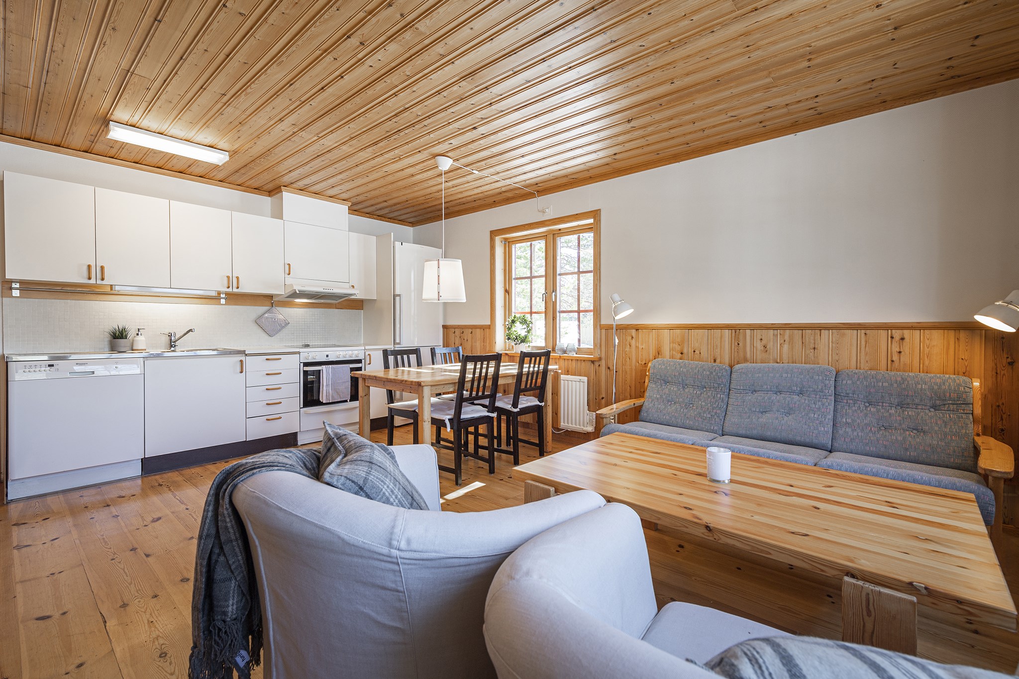 En hyggelig stue i en stil, der minder om Saalbach, med træloft og gulv. Det har lyse møbler, herunder en sofa, lænestole og et spisebord i træ med stole. Køkkenafdelingen er udstyret med hvide skabe og hårde hvidevarer. Et vindue lukker naturligt lys ind.