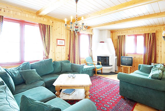 En hyggelig stue med trævægge og træloft, med grønne sofaer, en hvid pejs og et rødt mønstret tæppe. Et vintage-tv og en lysekrone, der minder om en Saalbach-skiferie, tilføjer et rustikt touch med naturligt lys, der strømmer ind gennem vinduerne.
