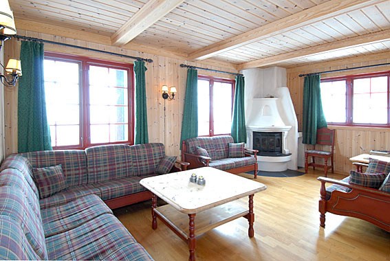 En hyggelig stue, der minder om en Saalbach skiferie, med trævægge og loftsbjælker. To plaidmønstrede sofaer og en lænestol omkranser et hvidt sofabord. I baggrunden varmer en hvid hjørnepejs rummet, mens grønne gardiner pryder vinduerne.