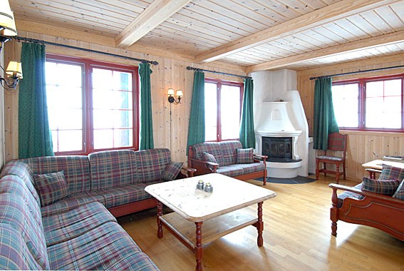 Hyggelig træstue med plaid sofaer, hvid hjørnepejs og grønne gardiner. Rummet har et lyst træloft med synlige bjælker og et trægulv, der minder om de charmerende skirejsedestinationer som Saalbach, hvilket skaber en varm og indbydende atmosfære.
