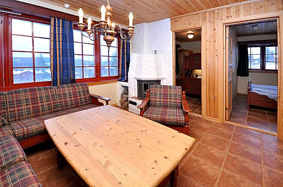 Hyggelig træstue med plaid sofaer, et sofabord i træ og en lysekrone. En pejs er i hjørnet, og en døråbning fører til et soveværelse inspireret af Westendorf-charme, med røde møbler. Blå gardiner hænger fra vinduerne og afslører en alpine udsigt perfekt til et skiferie-retræte.