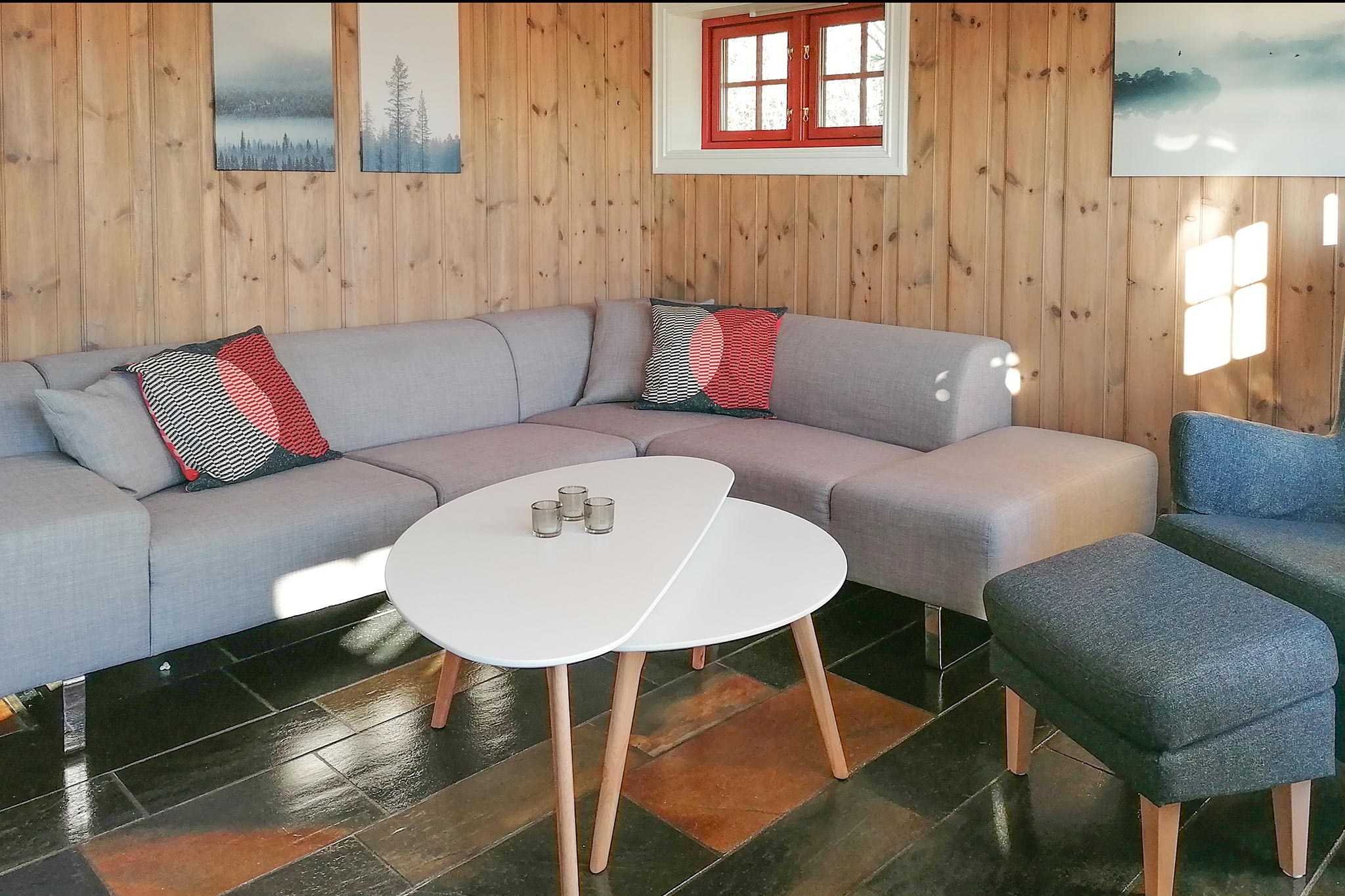 En hyggelig stue i Wagrain med en grå L-formet sofa prydet med røde og mønstrede hynder. Et lille hvidt sofabord og en mørkegrå stol hviler på et klinkegulv. Trævægge viser naturskønne kunstværker, der minder om skieventyr, mens et vindue lukker sollys ind.