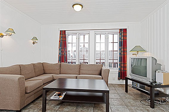 En hyggelig stue perfekt til afslapning efter en dag med skiferie, med en beige sofagruppe, et mørkt træ sofabord og et vintage-tv. Det veloplyste rum har store vinduer indrammet af farverige mønstrede gardiner og neutrale klinkegulve, der minder om et charmerende Saalbach-retræte.