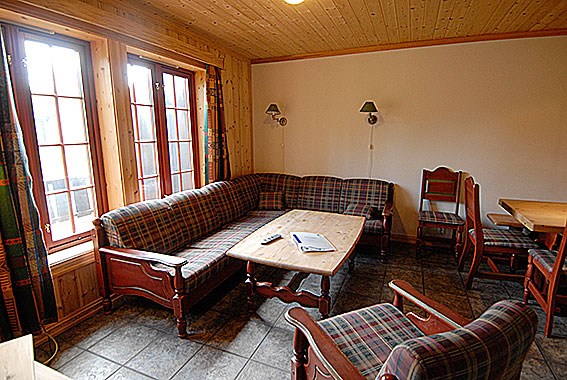 En hyggelig stue med plaidmøbler, herunder en hjørnesofa og to stole, der minder om en Saalbach-hytte. Et sofabord i træ rummer en fjernbetjening og et magasin, mens store vinduer med trærammer slipper naturligt lys ind. En spiseplads med stole er synlig til højre.