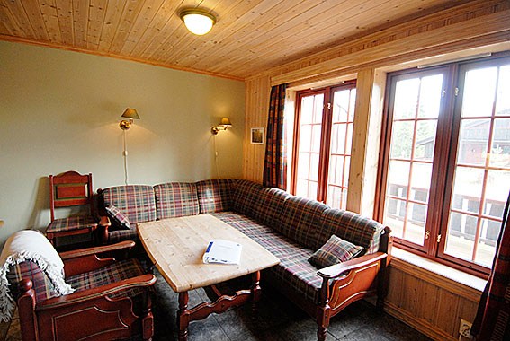 Et hyggeligt værelse med trævægge og -loft, med en plaidmønstret hjørnesofa og matchende lænestole omkring et træbord. To væglamper oplyser rummet, mens store vinduespartier giver naturligt lys og udsigt, der minder om en sommerferie Østrig-ferie.