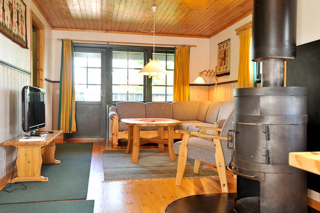 En hyggelig stue, der minder om et skiferie-retræte, med træloft og gulve, har en grå sektionssofa, et rundt sofabord og en sort brændeovn. Store vinduer med gule gardiner indrammer rummet som Saalbach-kulisser. Varm, rustik indretning væver sig gennem den dejlige opsætning.