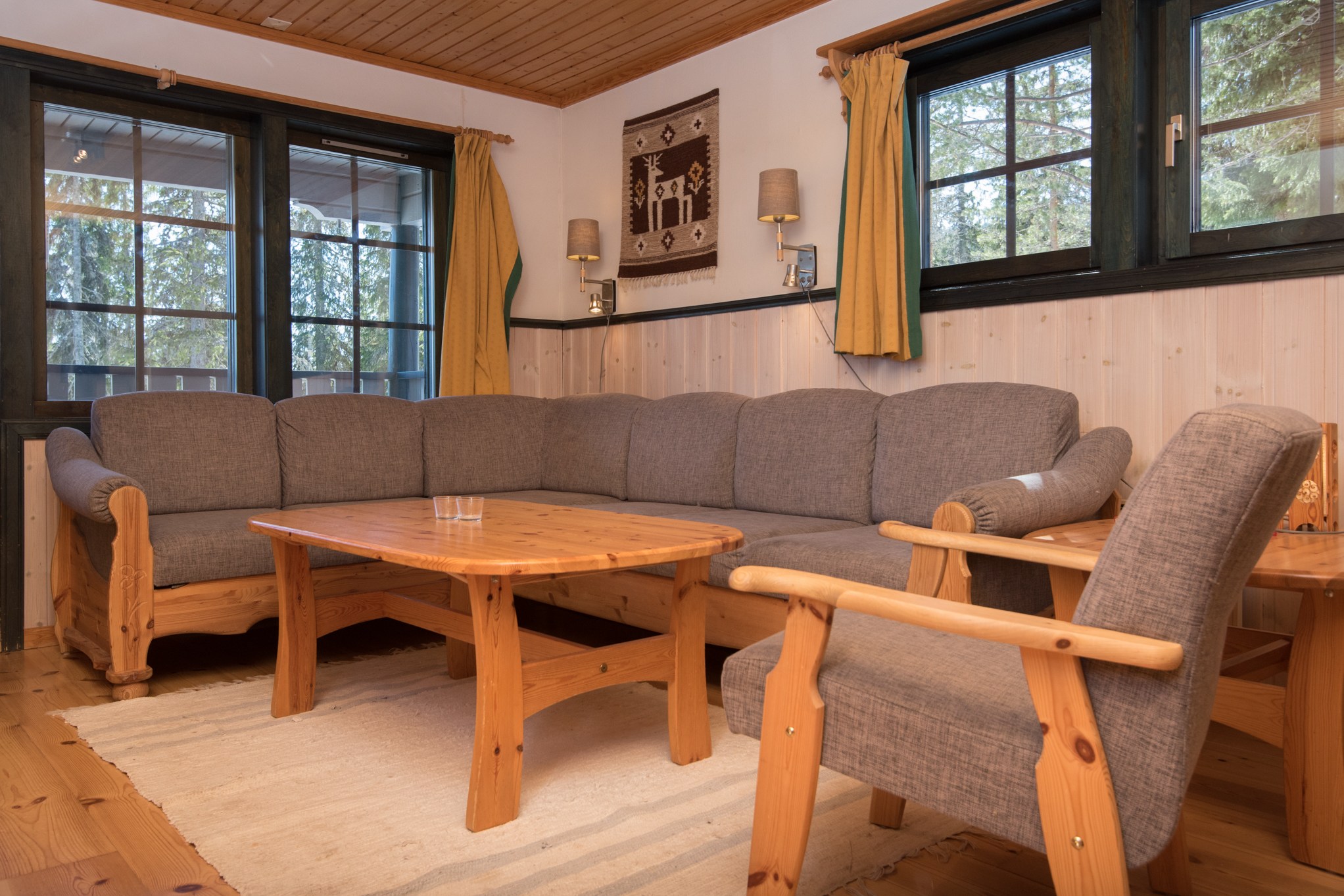 En hyggelig stue i Westendorf med trævægge og trægulv viser en grå L-formet sofa og matchende lænestol. Et sofabord i træ står på et lyst tæppe, mens gule gardiner indrammer vinduerne. Et dekorativt vægbeklædning tilføjer karakter over sofaen, der minder om et skiferie-retræte.