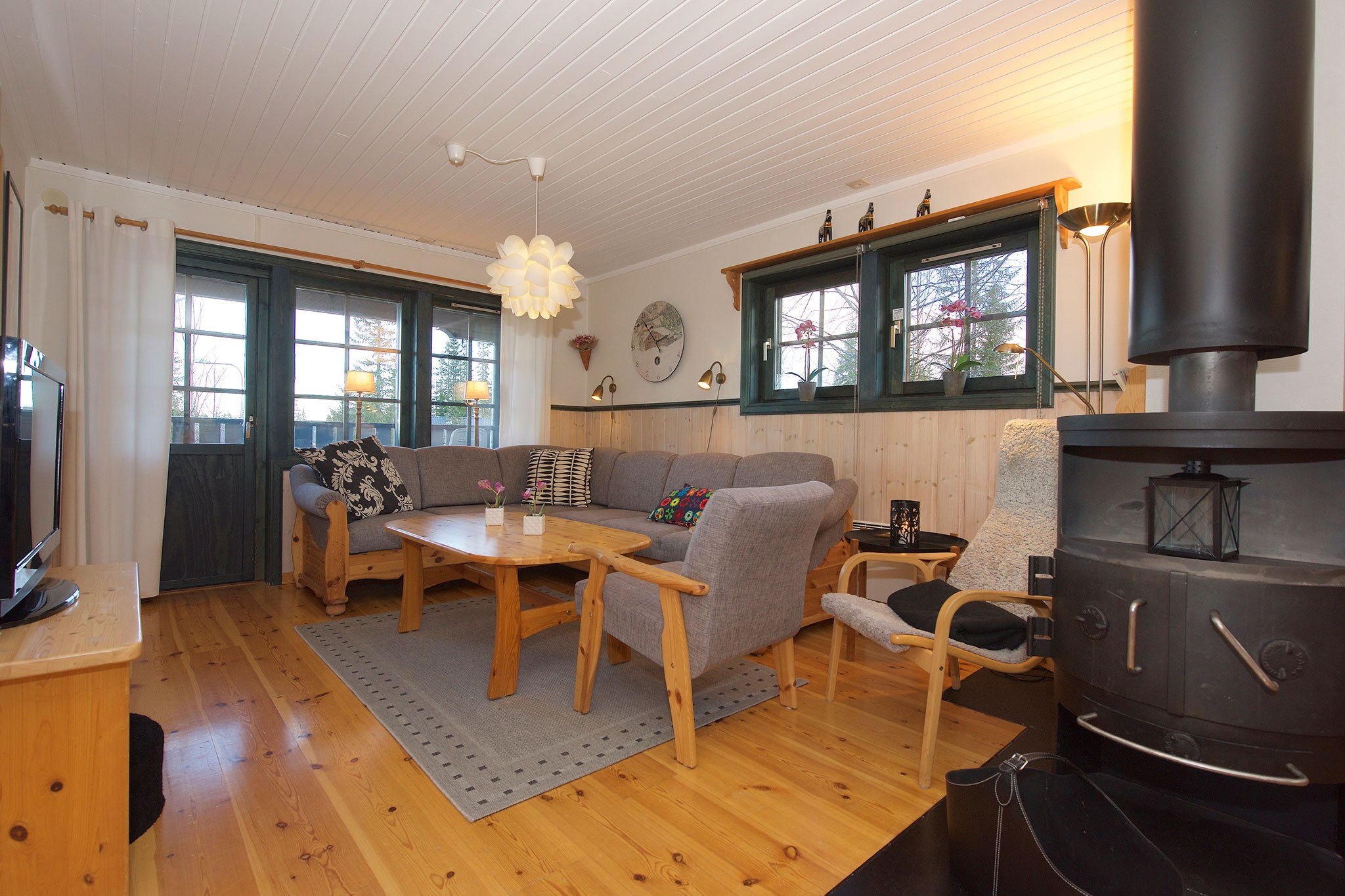 En hyggelig stue med trægulv og loft fremkalder charmen ved Westendorf. Den har en grå hjørnesofa, sofabord i træ, lænestol og en sort brændeovn. Store vinduespartier med grønne rammer slipper naturligt lys ind. Skandinaviske indretningselementer er til stede.