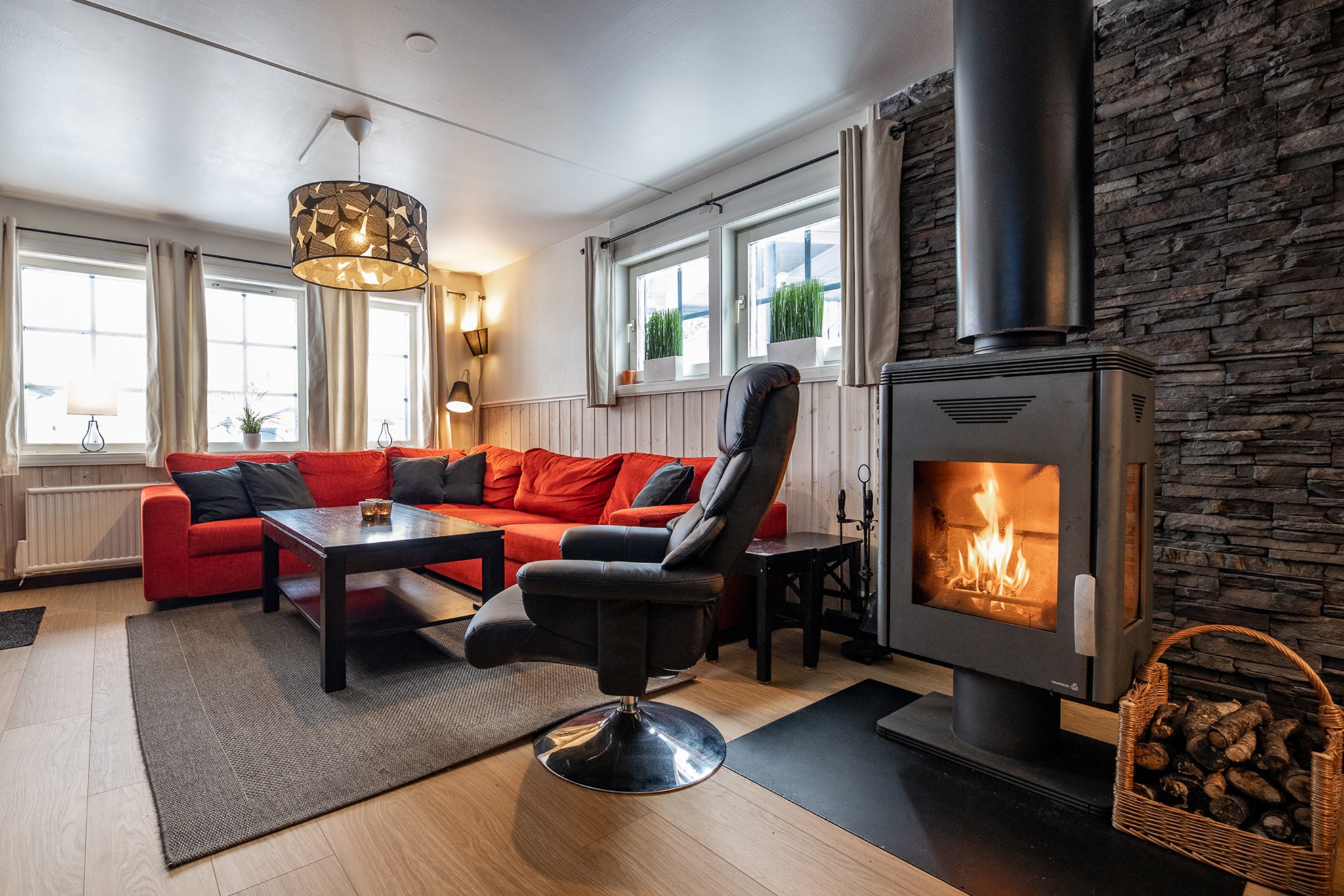 En hyggelig stue i Westendorf har en lys rød sofa, en sort hvilestol og en moderne brændeovn med ild. Der er en stenmur, store vinduer og et sofabord på et gråt tæppe, der minder om indbydende afterski-sammenkomster under skiferien.