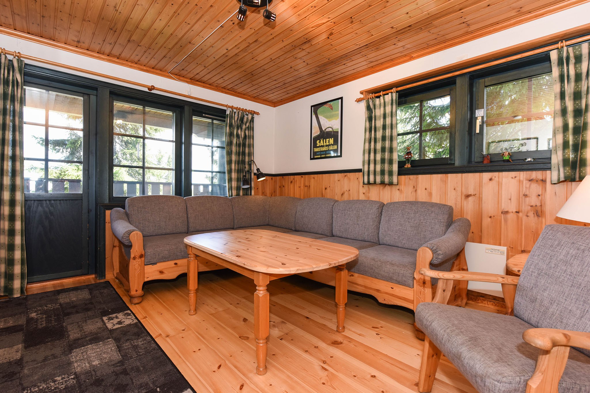 En hyggelig stue i Westendorf med hjørnesofa, sofabord i træ og lænestol. Værelset har rustikke trævægge, ternede gardiner og en naturskøn plakat. Store vinduer og en glasdør giver naturligt lys, perfekt til at nyde charmen ved dit skiferie-retræte.