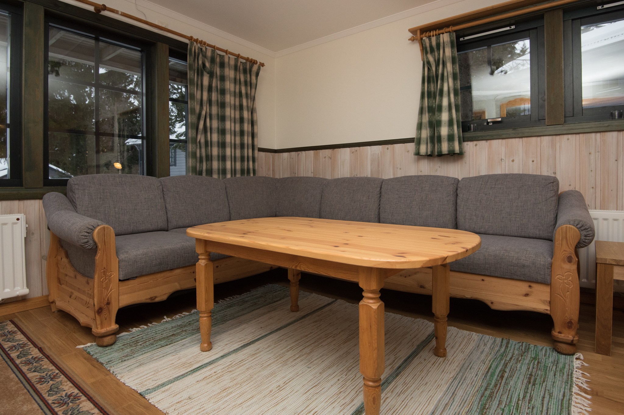 En hyggelig stue med et hjørnesofasæt i træ med grå hynder, der minder om en charmerende Westendorf-hytte. Et matchende sofabord i træ komplementerer de grønne plaidgardiner. Gulvet er udsmykket med et enkelt områdetæppe, og væggene kan prale af elegante træpaneler.