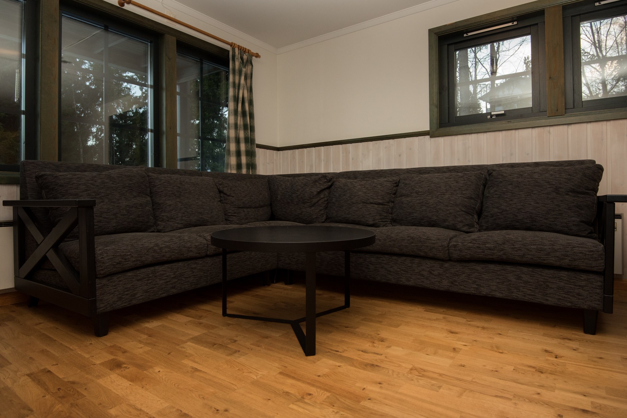 En stue i Saalbach har en mørkegrå L-formet sofa med et rundt sort sofabord foran. Trægulvet, vinduet med plaidgardiner og vægge med træpaneler skaber en hyggelig atmosfære, der minder om et sommerferie Østrig-retræte.