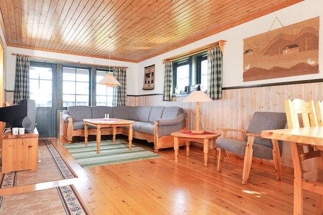 En hyggelig stue, der minder om et Westendorf-retræte med trægulve og loft. Den har en grå sofa, en lænestol, et sofabord i træ og et tv. Sollyset strømmer gennem store vinduer ind på et grønstribet tæppe og skaber en alpin stemning perfekt efter skirejseeventyr.