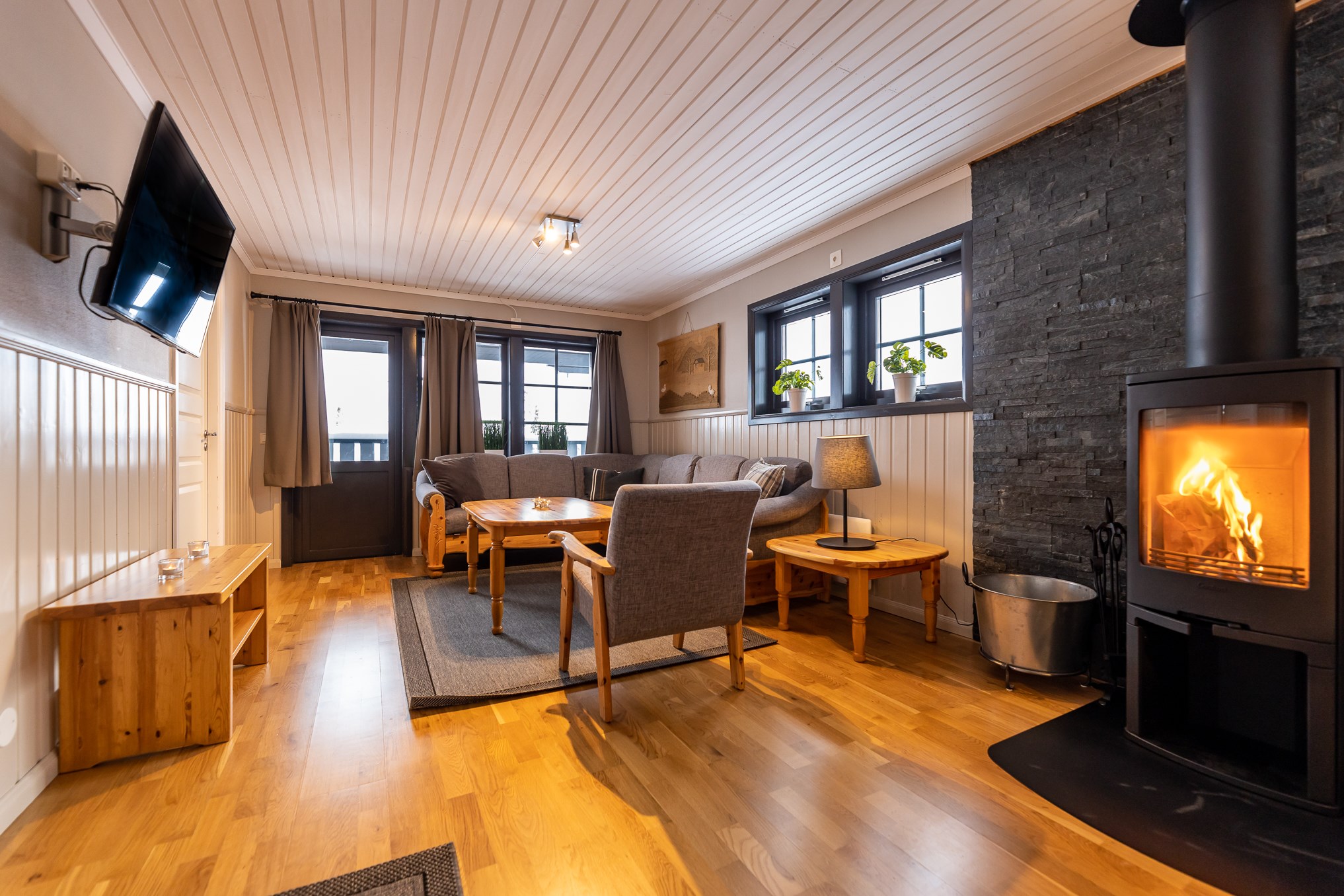 Hyggelig stue med trægulve, moderne pejs og komfortable grå siddepladser. Store vinduer giver naturligt lys, perfekt til afslapning efter en dag på skiferie i Westendorf. Et fladskærms-tv er monteret på væggen, mens et træbord og små indendørs planter tilføjer varme til rummet.