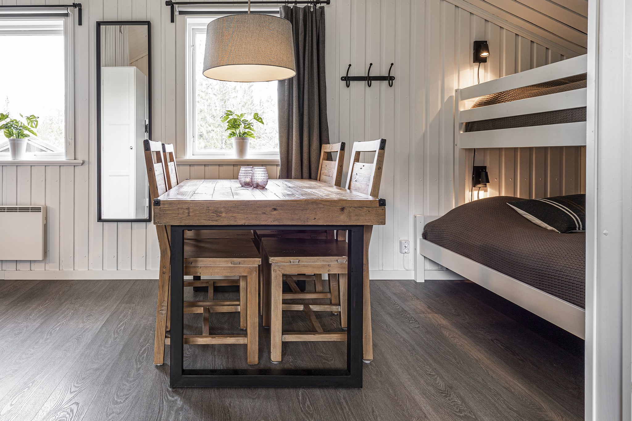 Hyggeligt værelse med et spisebord i træ og fire stole, placeret ved siden af et vindue prydet med planter. En hængende lampe oplyser rummet. Der er en køjeseng og et spejl i fuld længde på væggen, hvilket skaber en varm, hjemlig atmosfære perfekt til at slappe af efter en dag i Wagrain.