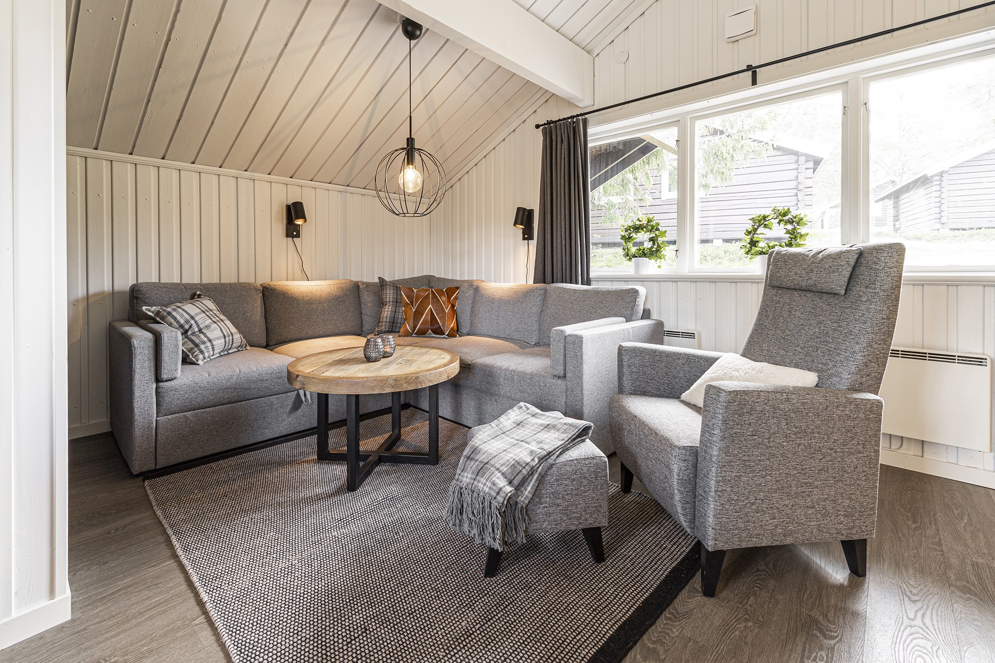 Hyggelig stue med en delt grå sofa, et rundt sofabord i træ og en matchende lænestol med skammel. Blød belysning forstærker varmen, der minder om sommeraftener i Westendorf. Neutral indretning inkluderer puder og et plaidkast, der tilbyder en indbydende ferielignende atmosfære.