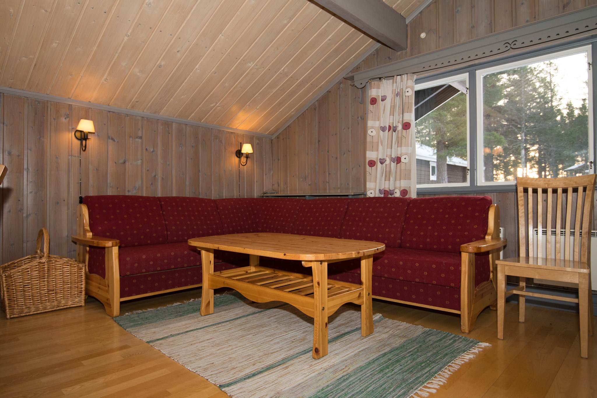 En hyggelig stue i stil med et Westendorf-retræte med trævægge og -loft, med en rød polstret L-formet sofa, et sofabord i træ og en vævet kurv. Et vindue med blomstergardiner lukker naturligt lys ind, mens et stribet tæppe dækker trægulvet.