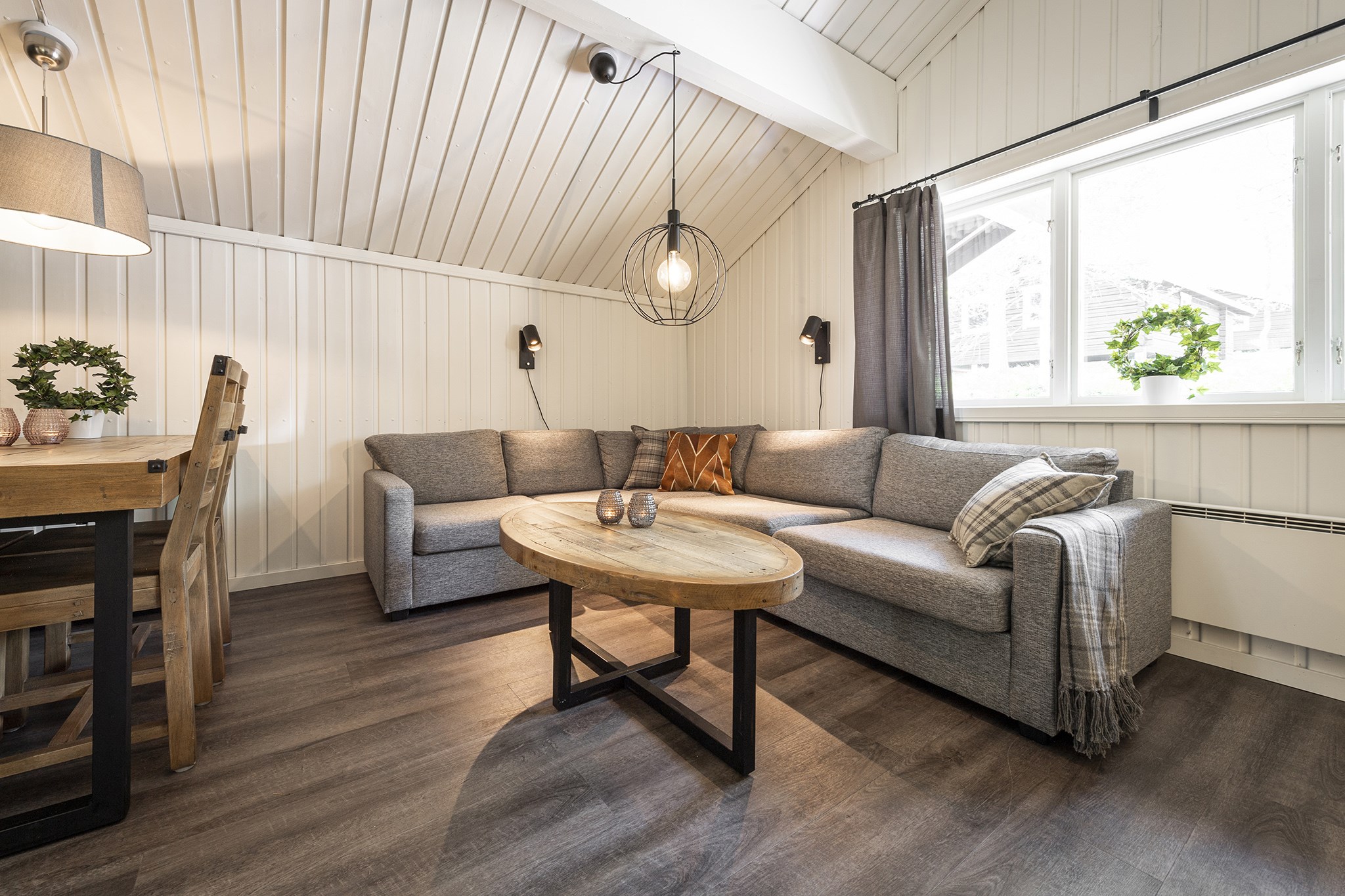 En hyggelig stue i en stil, der minder om Westendorf, med en grå sektionssofa, et ovalt sofabord i træ og et plaid-tæppe. Rummet har hvide panelvægge, loft til kip og stort vindue med grå gardiner. Et spisebord er synligt til venstre prydet med potteplanter.
