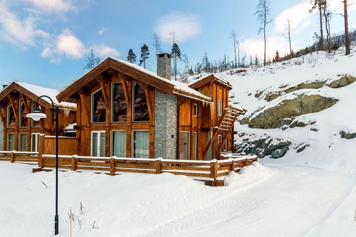 En træhytte med store vinduer og en stenskorsten ligger i et snelandskab, perfekt til en skiferie i Westendorf. Hytten er omgivet af træer og en klippeskråning og har et træhegn, der grænser op til den snedækkede sti, der fører til dens indbydende indgang.