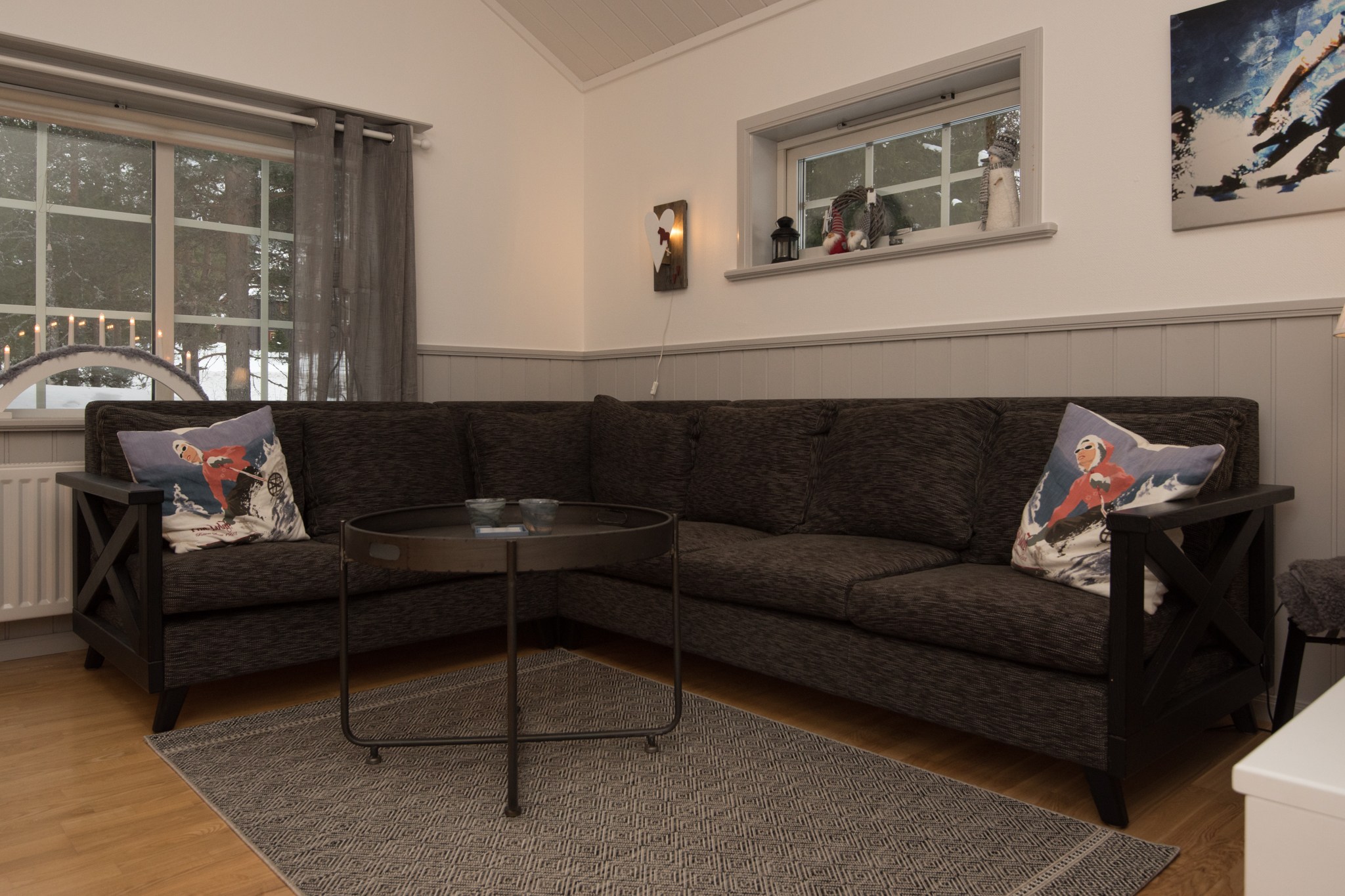 En hyggelig stue, der minder om et sommerferiested i Westendorf, har en mørkegrå hjørnesofa med to dekorative puder. Et rundt, sort sofabord sidder oven på et beige tæppe, mens rene gardiner tillader naturligt lys at oplyse vægkunst og stearinlys på skærmen.