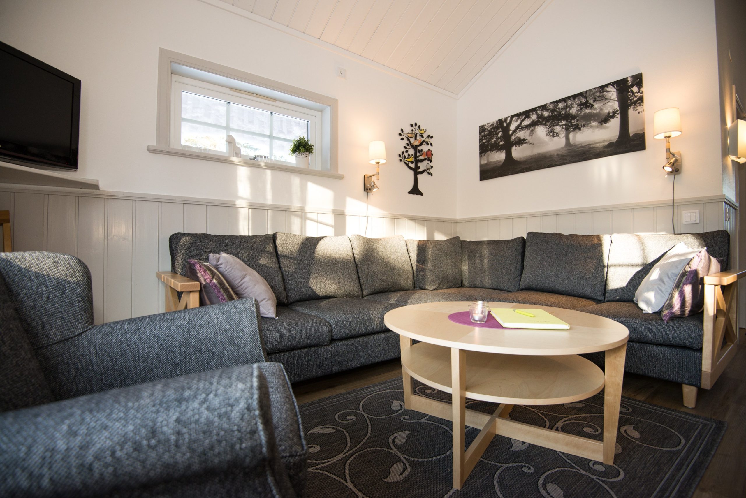 Hyggelig stue med grå sektionssofa, sofabord i træ og lænestol. Hvide vægge har et trækunstværk og sort-hvidt landskabsfoto, der minder om Saalbachs fredfyldte udsigter. Et tv er monteret i hjørnet, mens et lille vindue lader sollys strømme ind som en perfekt dag på skirejser.