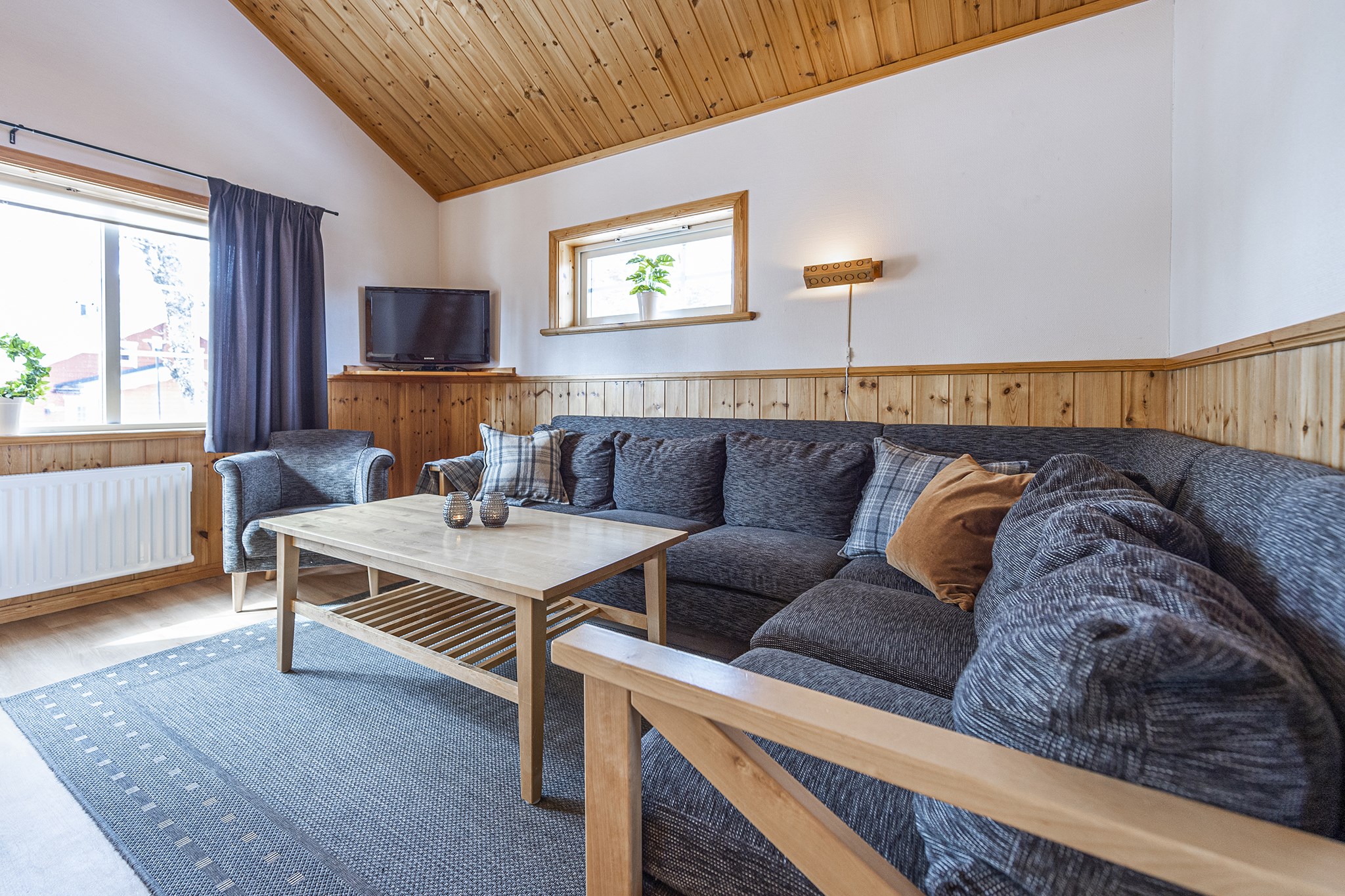 En hyggelig stue i Westendorf har en stor grå sektionssofa, sofabord i træ og hynder. Værelset kan prale af træpanelvægge, højt træloft, et lille tv og et vindue med gardiner og potteplanter, perfekt til afslapning efter en eventyrlig skiferie.
