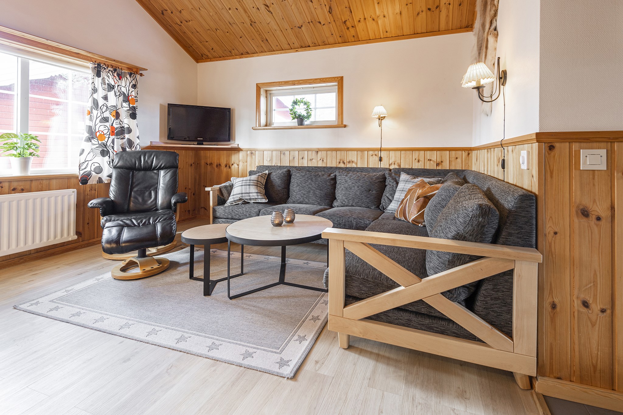 En hyggelig stue med trævægge og loft fremkalder charmen ved et skirejser-retreat. Den har en mørkegrå sektionssofa med hynder, en sort drejestol, runde sofaborde, et vægmonteret tv og et vindue med mønstrede gardiner. Et lille tæppe med stjernedesign ligger på gulvet.