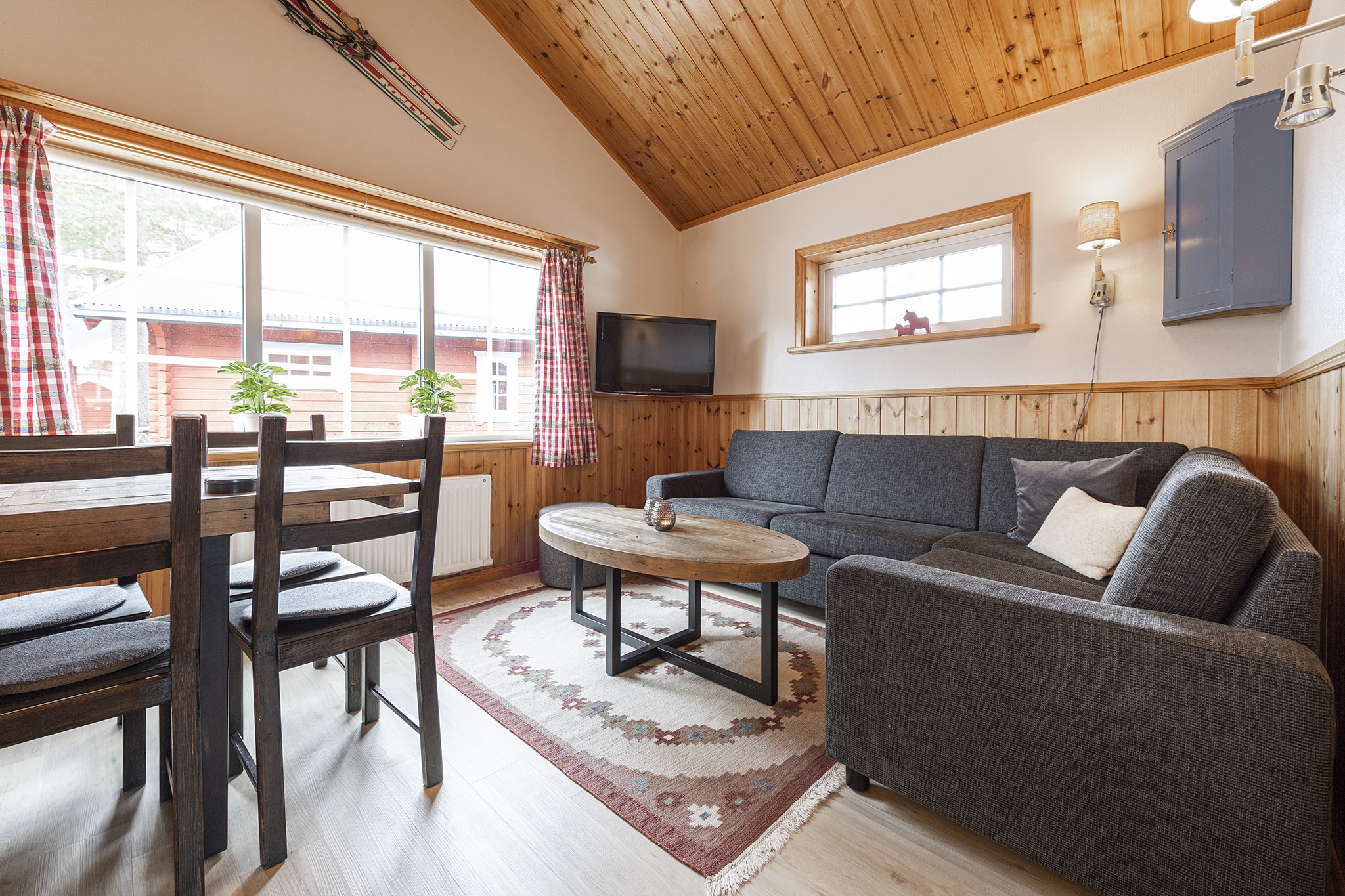 En hyggelig stue i Saalbach med trævægge og loft. Den har en grå sektionssofa, et rundt sofabord i træ, et spisebord med fire stole og et vægmonteret tv. Store vinduer med plaidgardiner lyser op i rummet efter skirejsers eventyr.