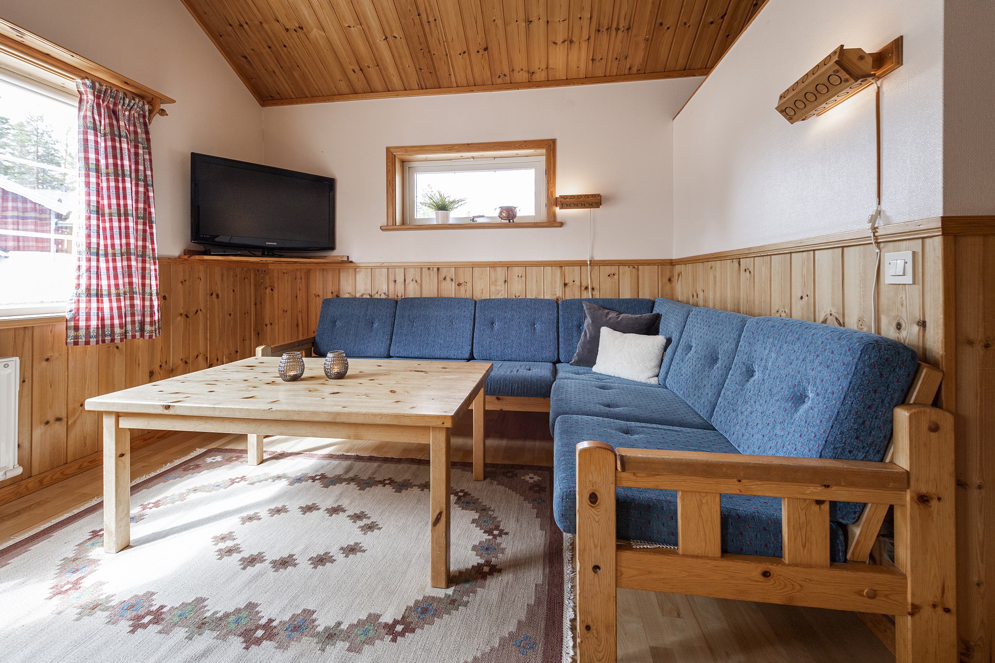 En hyggelig stue med træpaneler på vægge og loft, der minder om et Saalbach-retræte, med en blå L-formet sofa, et sofabord i træ og et tv i hjørnet. Et mønstret tæppe pryder gulvet, mens sollys filtreres gennem plaidgardiner. Perfekt til at planlægge din sommerferie Østrig.