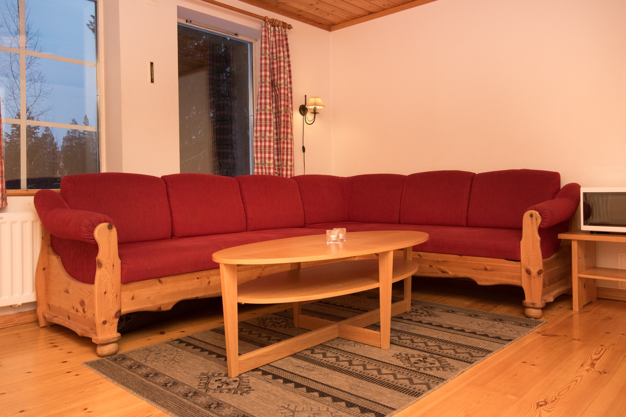 En hyggelig stue i stil med Saalbach, med en rød L-formet sofa sat mod trævægge, der minder om Westendorfs alpine charme. Et sofabord af træ hviler på et mønstret tæppe, mens plaidgardiner indrammer vinduet og lukker naturligt lys ind, der forstærker dette sommerferie Østrig-tilflugtssted.