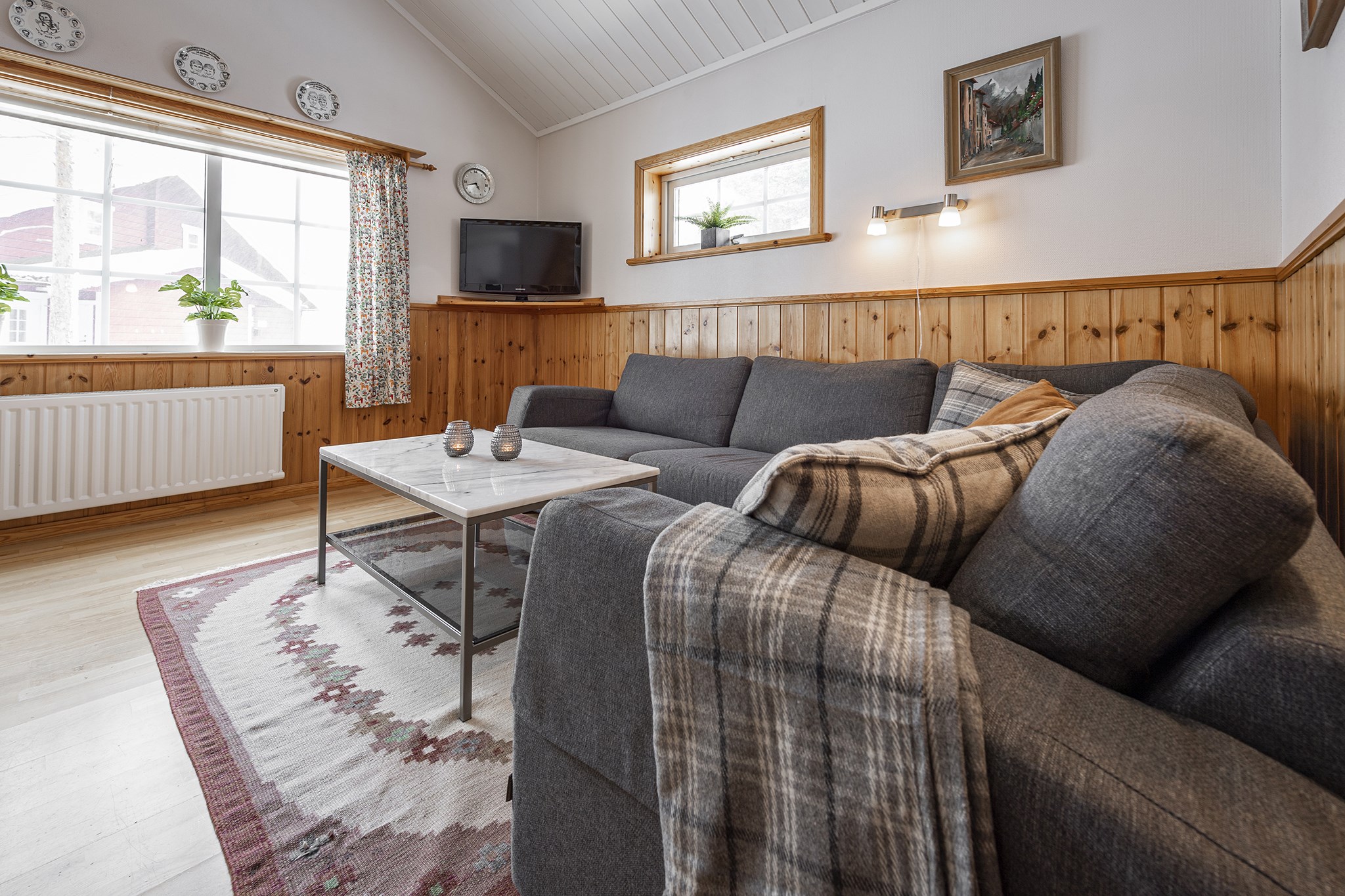 En hyggelig stue, der minder om en Saalbach skiferie-hytte, har en grå sektionssofa med plaid, et marmor sofabord og et tæppe. Vægge med træpaneler er prydet med plader og kunst. Store vinduespartier slipper naturligt lys ind, og et tv er placeret i hjørnet.