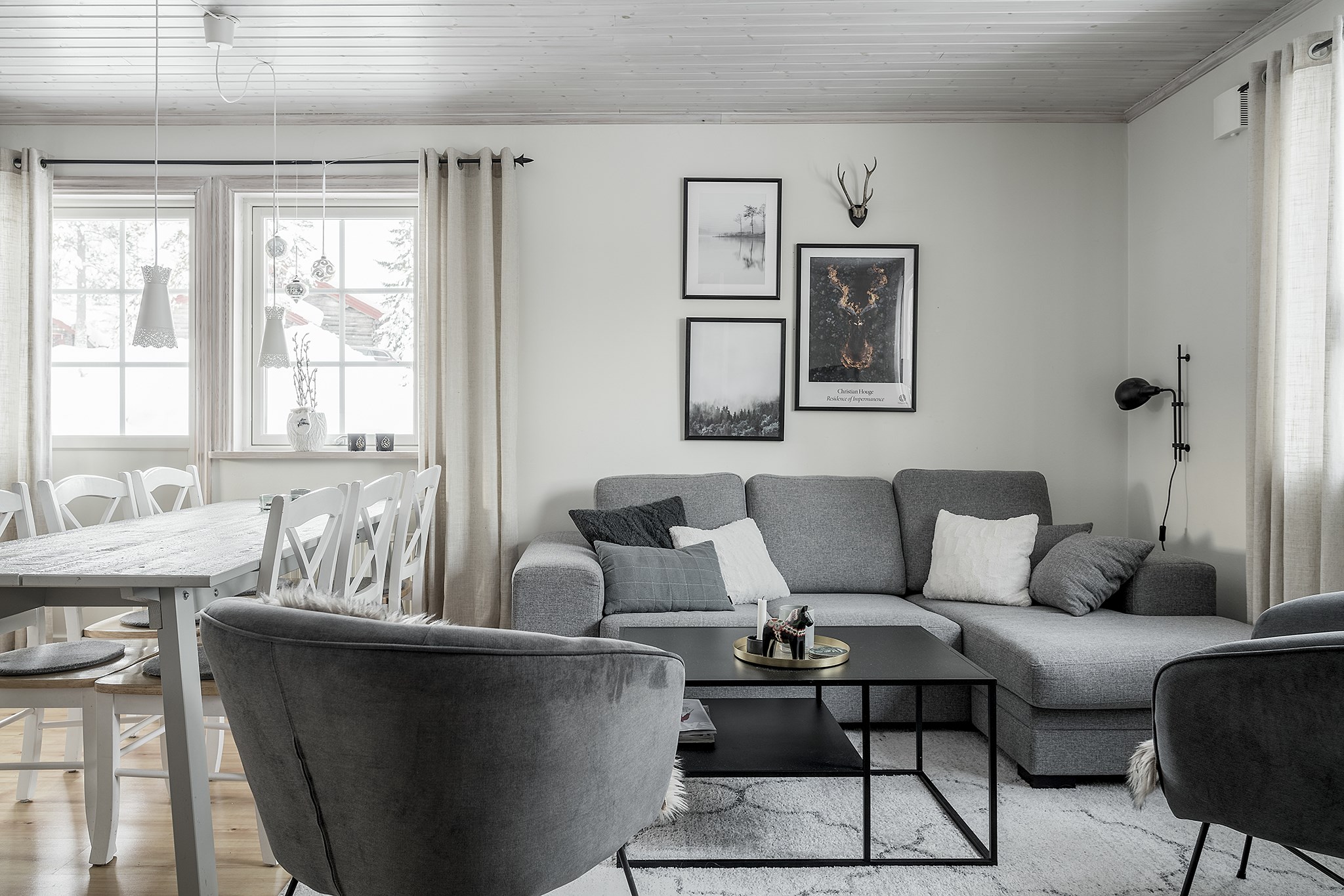 En hyggelig stue, der minder om et Saalbach-retræte, har en grå sektionssofa, sort sofabord og to grå lænestole. Indrammede billeder og en hjortedekoration hænger over sofaen. Et spisebord med hvide stole sidder tæt ved et stort vindue, med beige gardiner, der elegant indrammer rummet.