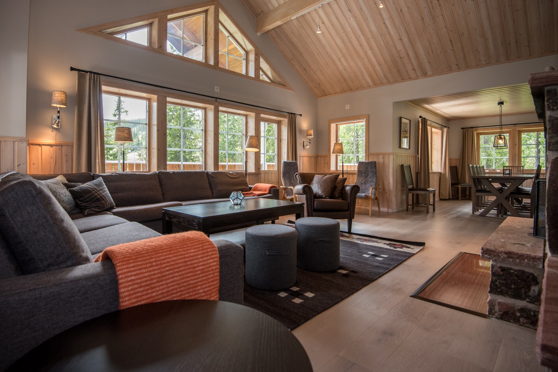 En hyggelig, moderne stue i Wagrain har en stor sektionssofa med et orange tæppe, to lænestole og et sofabord. Rummet er godt oplyst med store vinduespartier og højt til loftet i træ. En spiseplads er synlig i baggrunden, perfekt til at nyde skirejsehistorier.