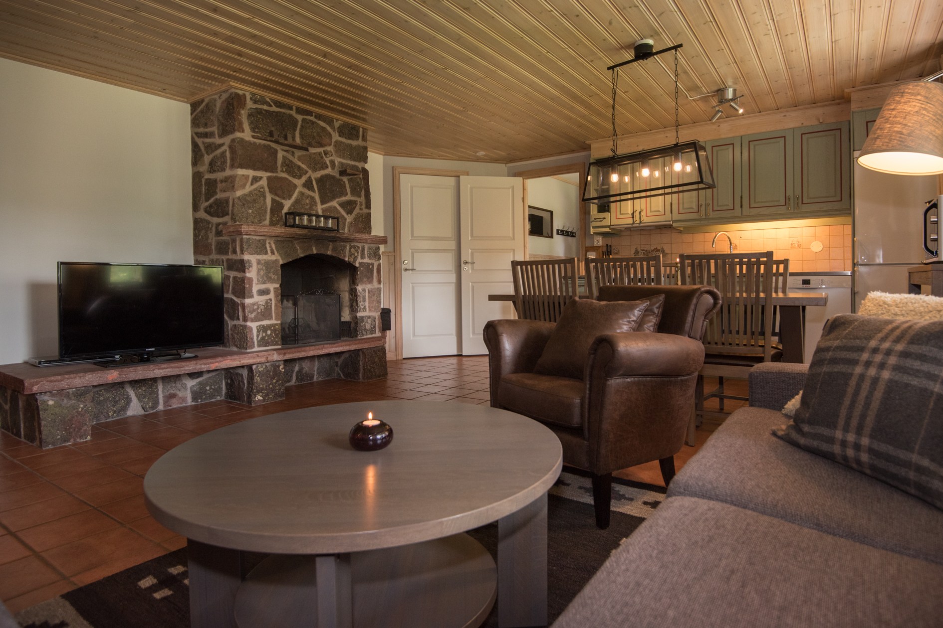 Hyggelig stue med stenpejs og fladskærms-tv, perfekt efter en dag i Saalbach. En brun lænestol, grå sofa og rundt sofabord med et tændt lys forstærker varmen. Værelset har træloft og åbent køkken med spiseplads i baggrunden.