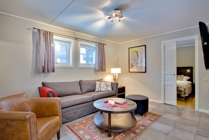 En hyggelig stue med en grå sofa, rød pude, brun læderstol, rundt sofabord og skammel udstråler varme. To vinduer med gardiner lyser rummet op som et Westendorf-retræte. En lampe og indrammet kunst tilføjer indretning, mens en døråbning afslører en del af et soveværelse.