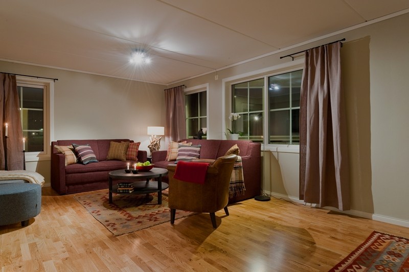 En hyggelig stue, der minder om et Westendorf-retræte, med en hjørnesofa, lænestol og rundt sofabord på et trægulv. Blødt oplyst med en lampe har rummet gardiner, der indrammer vinduerne. Et mønstret tæppe tilføjer varme til rummet, mens hynder er pænt arrangeret på sofaen.