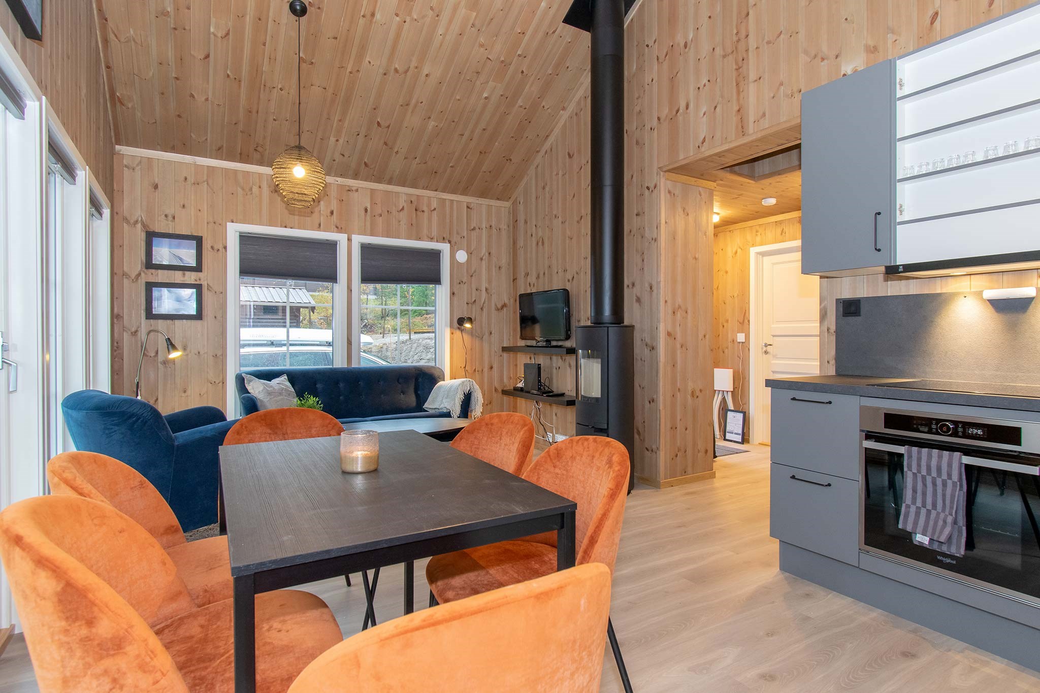 Et hyggeligt opholdsområde i åbent plan med trævægge og loft, perfekt til afterski-afslapning. Det har et orange spisebord, en blå sofa, en sort brændeovn og et moderne køkken. Store vinduespartier slipper naturligt lys ind og fremhæver den varme, indbydende indretning, der minder om Westendorf skiferie charme.