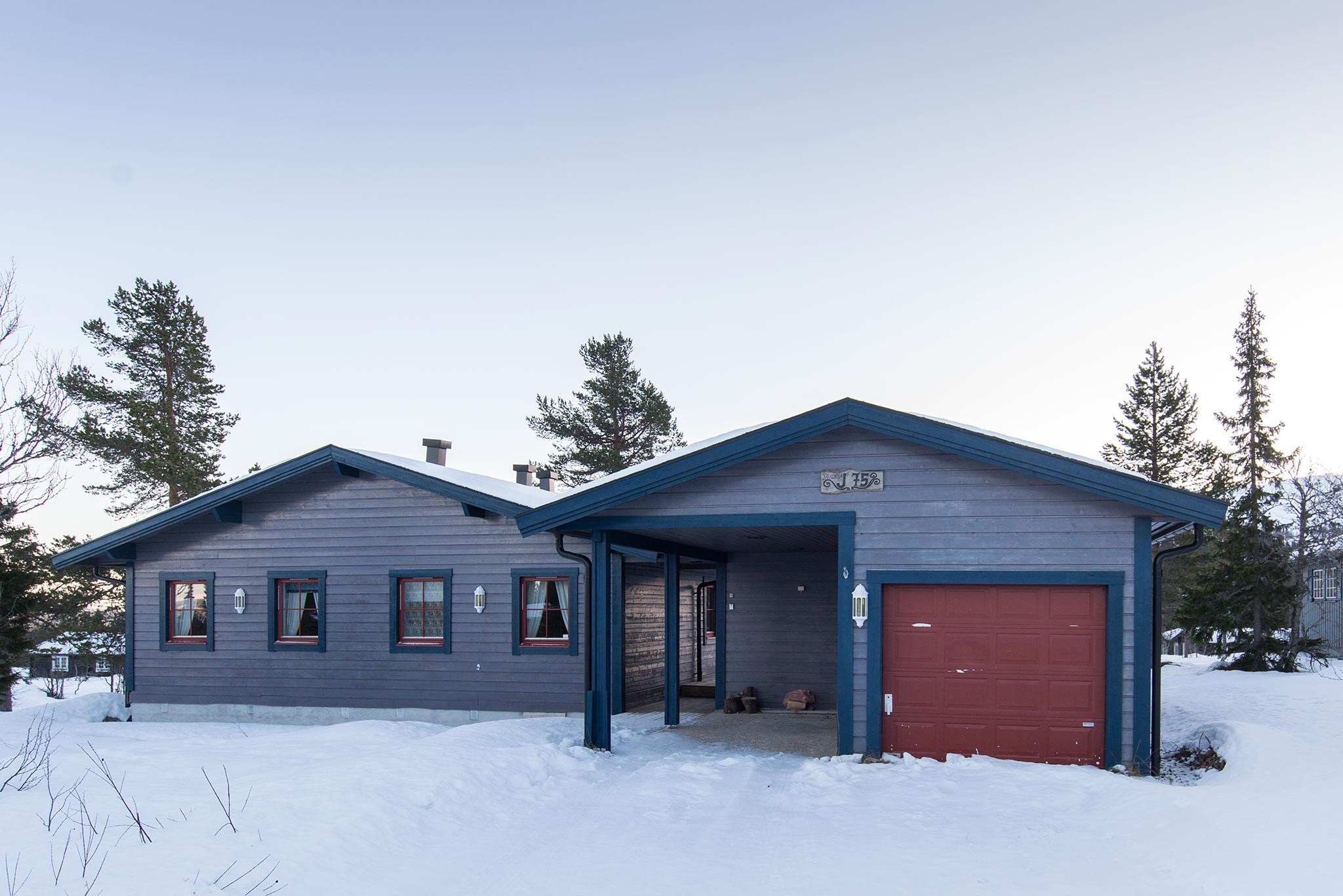 Et blågråt hus med en rød garageport ligger i et snedækket landskab, der minder om vinterdage i Wagrain. Flere vinduer med sorte rammer og en lille overdækket veranda tilføjer charme, mens fyrretræer pryder baggrunden under en klar himmel.