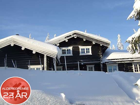 En snedækket træhytte under en klar blå himmel i det fredfyldte Westendorf. Taget er tungt belastet med sne. I forgrunden ses et skilt med Aldersgrense 23 år i rødt. Træer indhyllet i sne er synlige rundt om hytten, hvilket skaber en malerisk vinterscene.