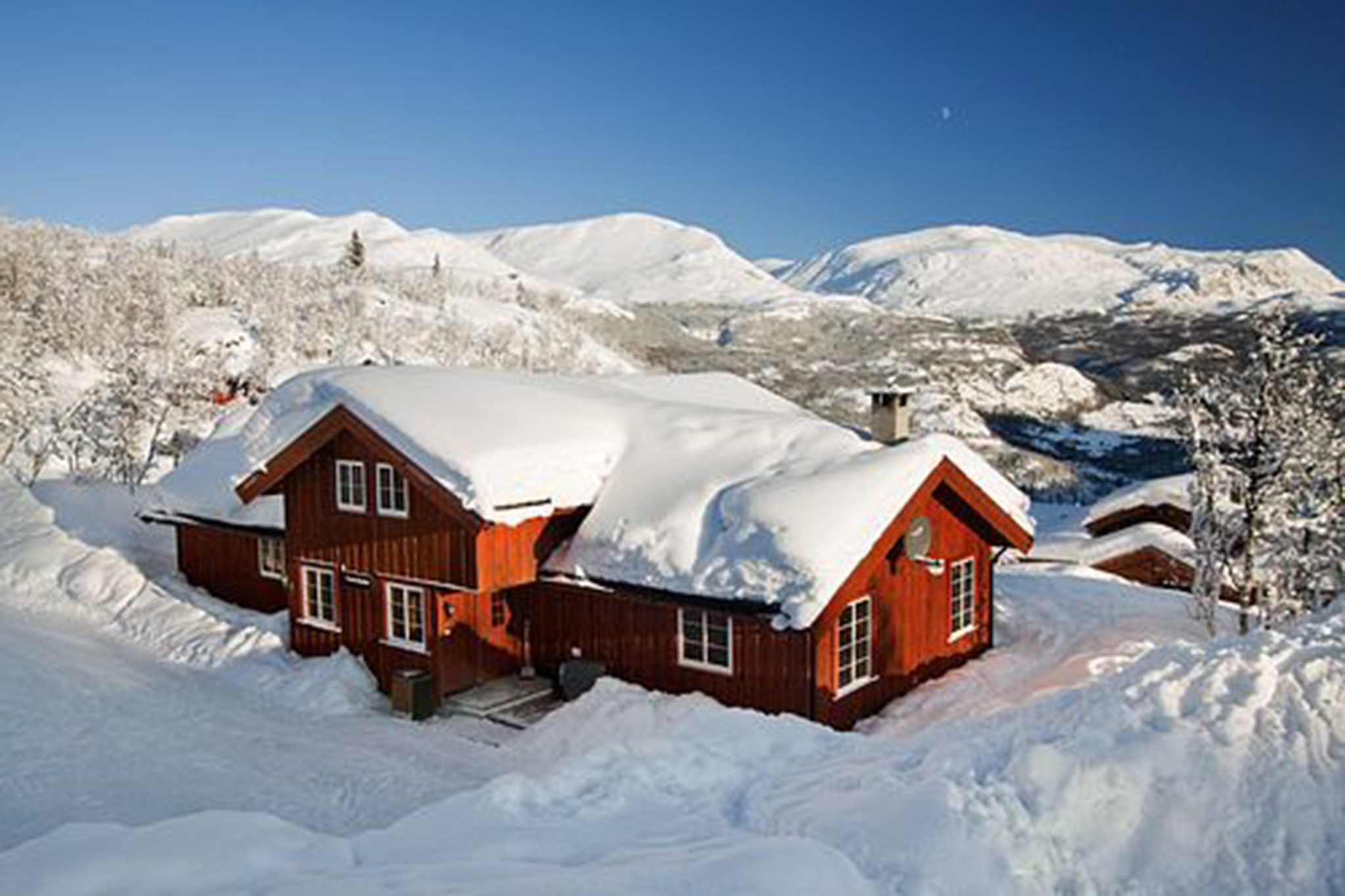 En rød træhytte omgivet af dyb sne i Wagrain, beliggende mod snedækkede bjerge under en klar blå himmel. Det delvist snedækkede tag og skorstenen fremkalder en rolig vinterlig charme, perfekt til en fredfyldt skiferieflugt.