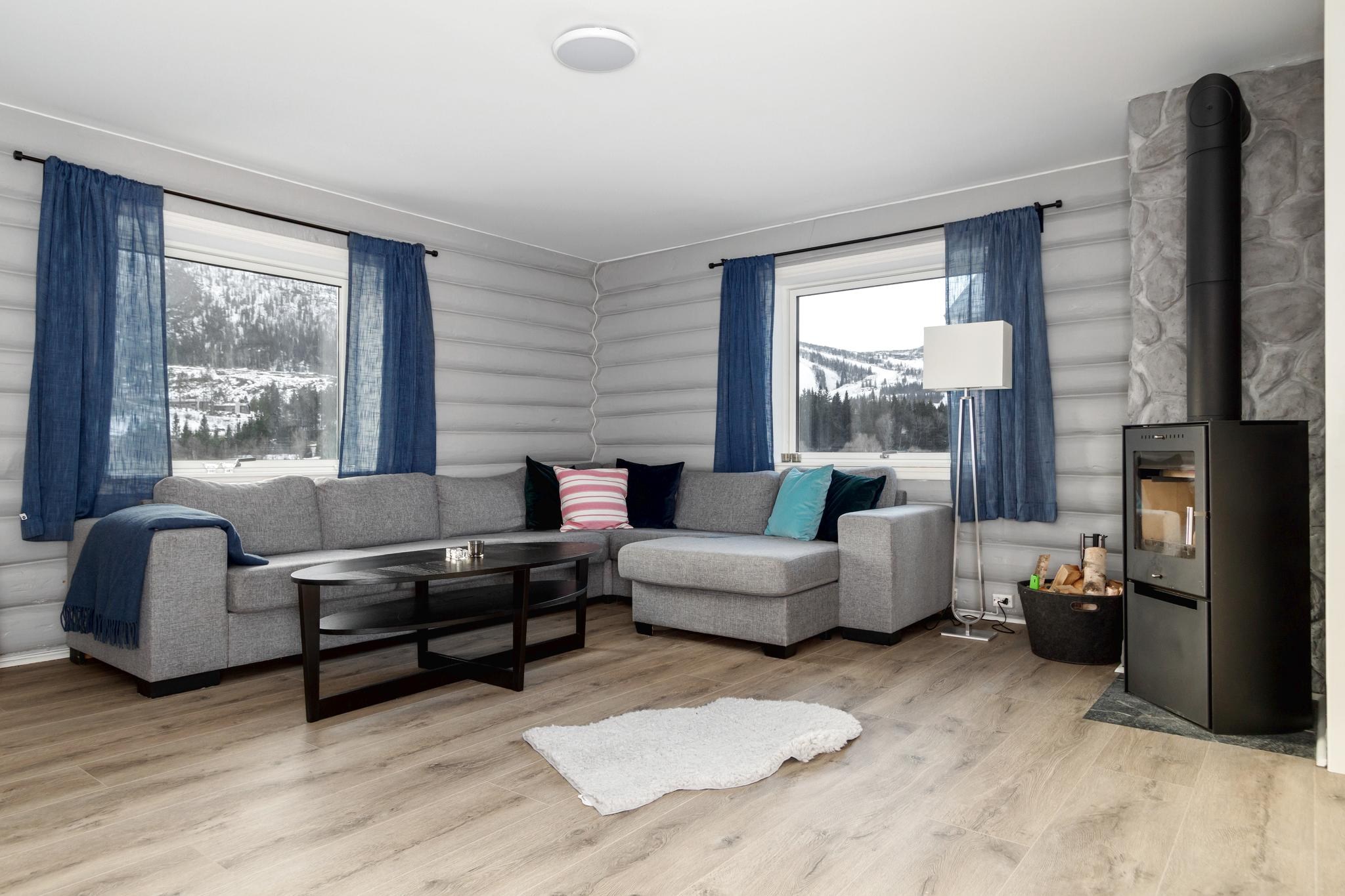 En hyggelig stue i Wagrain har en grå hjørnesofa, farverige hynder og et sort sofabord. Store vinduer med blå gardiner tilbyder en sneklædt udsigt over bjergene. En moderne brændeovn og et hvidt tæppe fuldender rummets varme atmosfære.